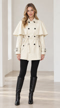 Front full-length view of woman wearing Cream capelet double-breasted trench coat with belt and knee-high boots